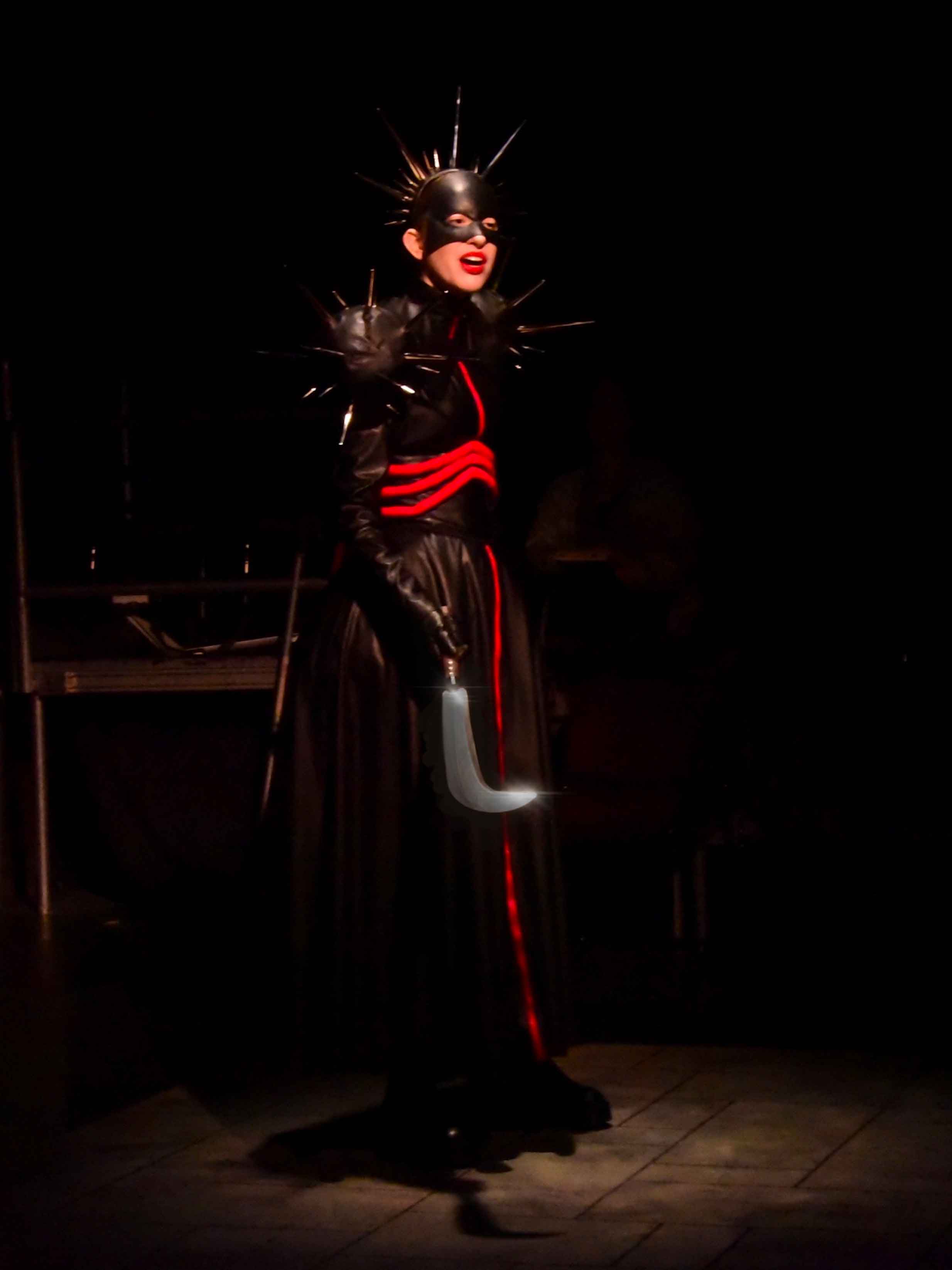 Actor wearing black dress with red detailing and sharp spikes coming out of the shoulders with a black mask and headpiece with sharp spikes coming out of the top of the head