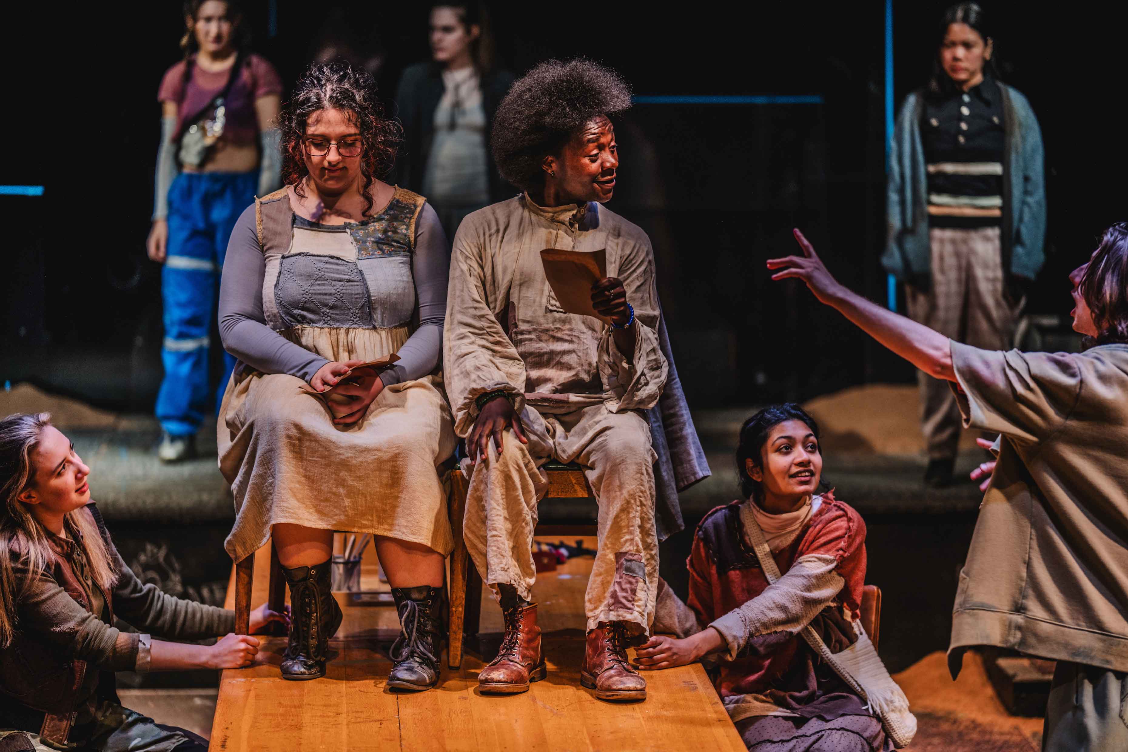 Two actors sit in chairs on table, while surrounding actors sit or stand nearby from scene in The Trials