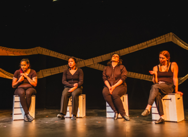 four women sit on boxes on stage with measuring tape on wall in background