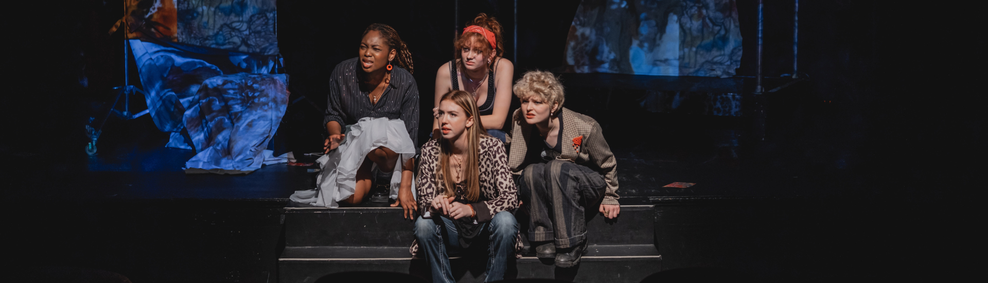 Four female actors sit on edge of stage looking off at something in the distance 